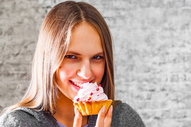 portrait of young teenager brunette girl with long hair eating cream cake on gray wall background - Фото, зображення