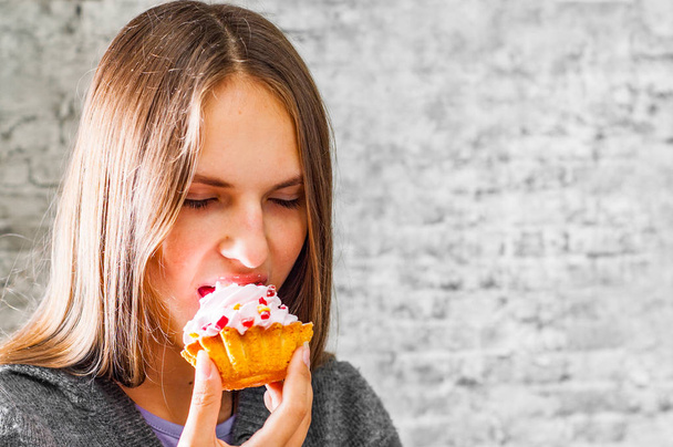 portrait of young teenager brunette girl with long hair eating cream cake on gray wall background - Foto, immagini