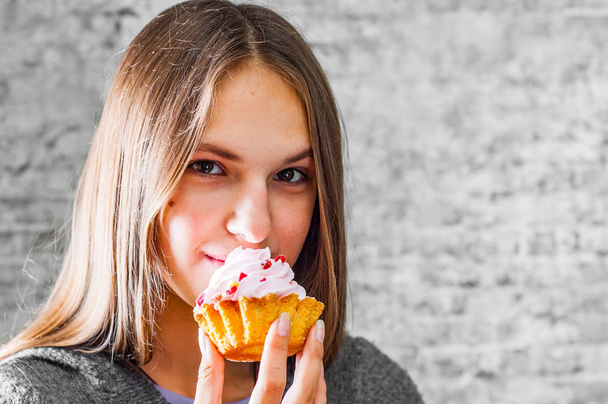 portrait of young teenager brunette girl with long hair eating cream cake on gray wall background - Фото, изображение
