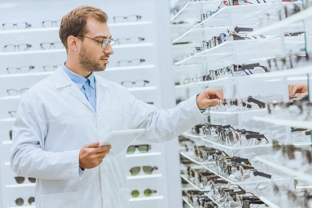 professional optician with tablet taking glasses from shelves in optica - Photo, Image