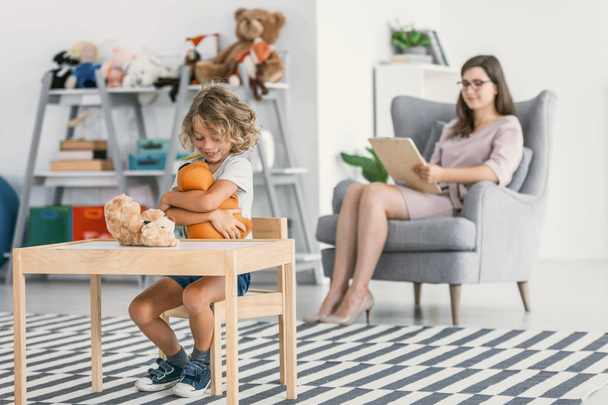 Happy autistic boy playing with a plush toy and his psychologist sitting in the blurred background holding a notepad - Фото, зображення