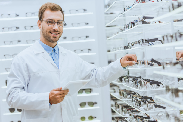 professional optometrist with digital tablet taking eyeglasses from shelves in optica - Photo, Image