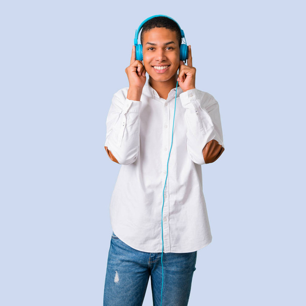 Jeune homme afro-américain avec chemise blanche écoutant de la musique avec écouteurs sur fond bleu isolé
 - Photo, image