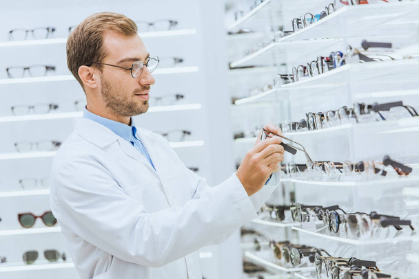 professional male oculist taking eyeglasses from shelves in ophthalmic shop - Foto, immagini