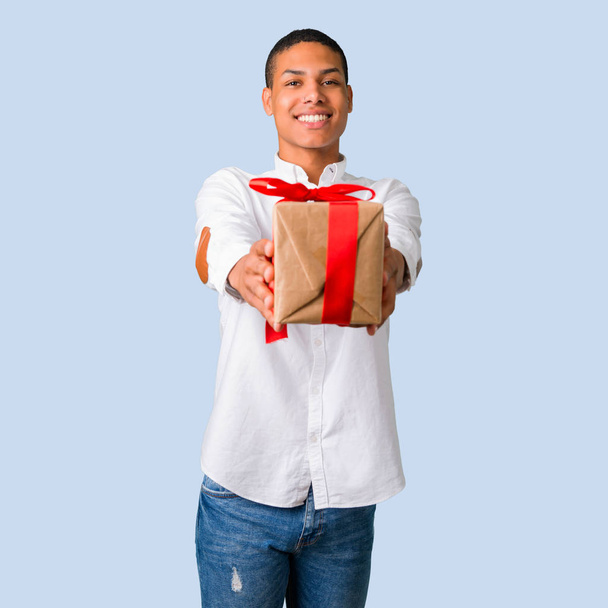 Jeune homme afro-américain avec chemise blanche tenant des boîtes-cadeaux dans les mains sur fond bleu isolé
 - Photo, image