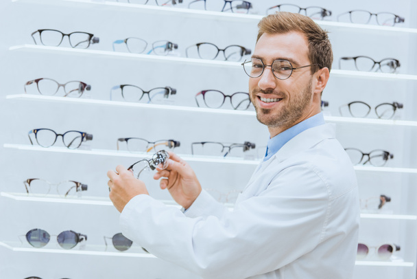 smiling doctor in white coat taking eyeglasses from shelves in optica - Photo, Image