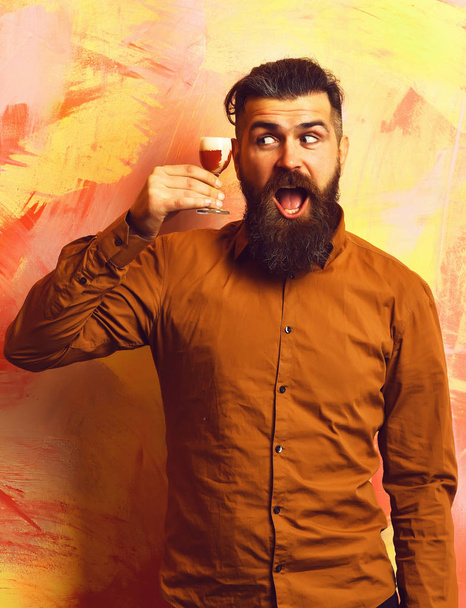 Bearded man, long beard. Brutal caucasian funny happy hipster with moustache in brown shirt holding alcoholic red shot on colorful texture background. - Photo, image
