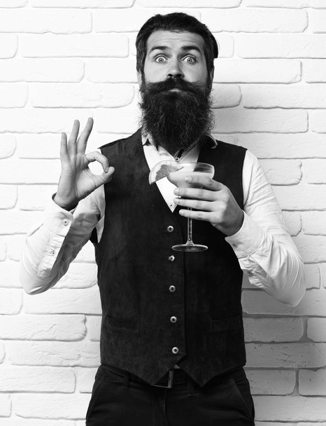handsome bearded man with long beard and mustache has stylish hair on surprised face holding glass of alcoholic beverage in vintage suede leather waistcoat o white brick wall studio background - Valokuva, kuva