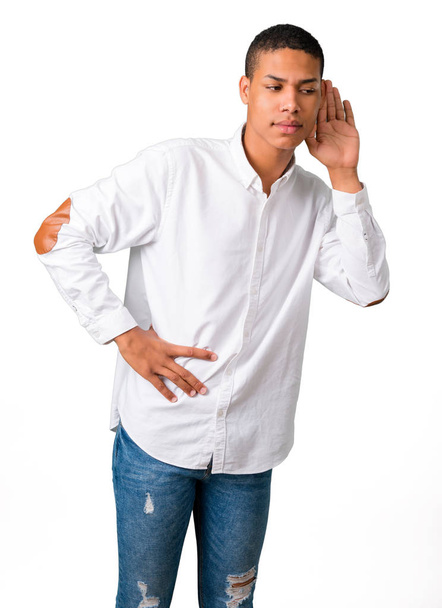 Jeune homme afro-américain avec chemise blanche écoutant quelque chose en mettant la main sur l'oreille sur fond blanc isolé
 - Photo, image