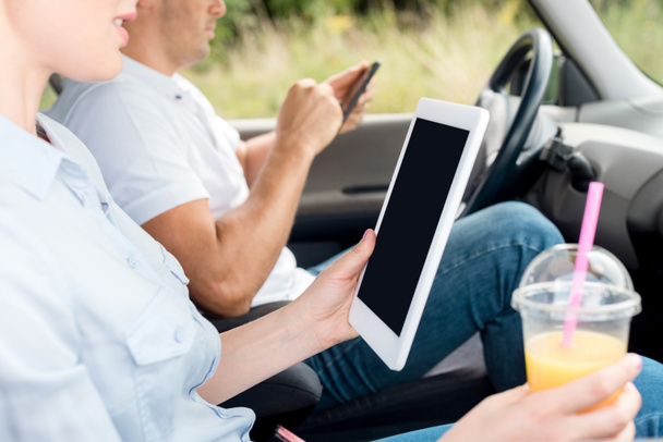 colpo ritagliato di donna con tazza di plastica di succo d'arancia utilizzando tablet in auto mentre l'uomo utilizzando smartphone sullo sfondo
 - Foto, immagini