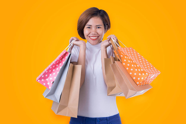 Portréja egy izgatott gyönyörű ázsiai lány viselt ruha gazdaság bevásárló táskák elszigetelt narancssárga háttér - Fotó, kép