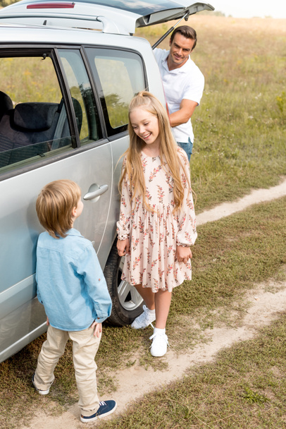 entzückende kleine Kinder, die neben dem Auto stehen, während ihr Vater während der Fahrt den Kofferraum im Feld öffnet - Foto, Bild