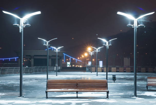 Πάγκος σε μια χιονισμένη πλατεία φωτίζεται από λάμπες με ανακλαστήρες. - Φωτογραφία, εικόνα