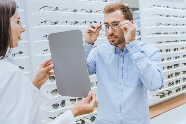 vista laterale dell'optometrista femminile che tiene lo specchio mentre l'uomo sceglie gli occhiali in ottica
  - Foto, immagini