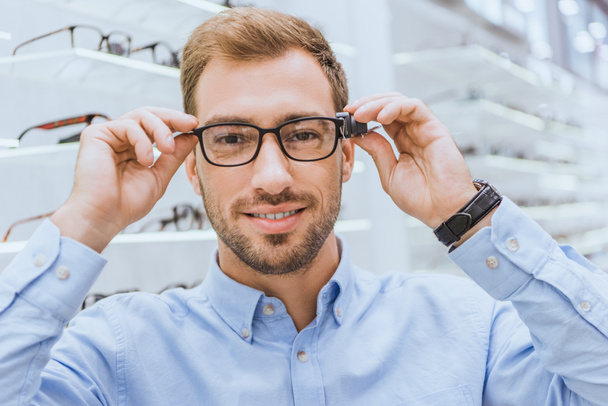 portrait of young man choosing eyeglasses and looking at camera in optics  - Fotoğraf, Görsel