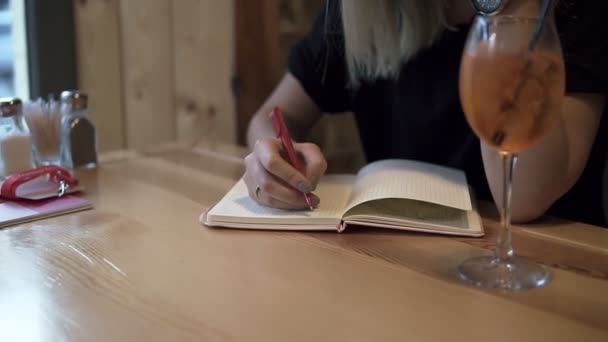 Mujer rubia de pelo largo sentada en un café moderno y tomando cuidadosamente algunas notas en su cuaderno. Un vaso de cóctel con hielo sobre la mesa. De cerca.
 - Imágenes, Vídeo