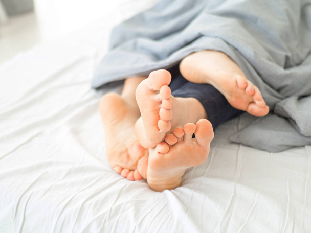 Voeten van het paar slapen naast elkaar in een comfortabel bed. Close up van voeten in een bed onder een witte deken. Blote voeten van een man en een vrouw gluren uit onder de cove. - Foto, afbeelding