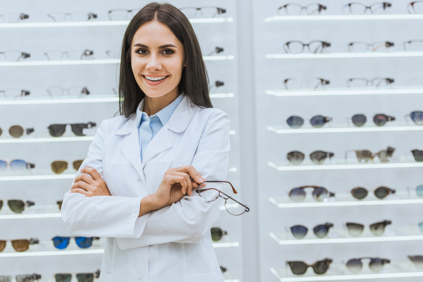 professioneller lächelnder Optiker mit Brille in der Nähe von Regalen in Optik - Foto, Bild