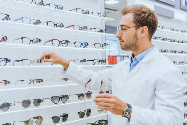 professional oculist taking eyeglasses from shelves in ophthalmic shop - Foto, Bild