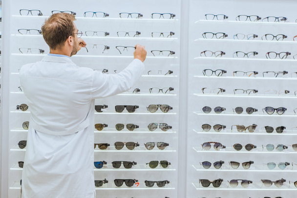 back view of oculist in white coat taking glasses from shelves in ophthalmic shop - Fotó, kép