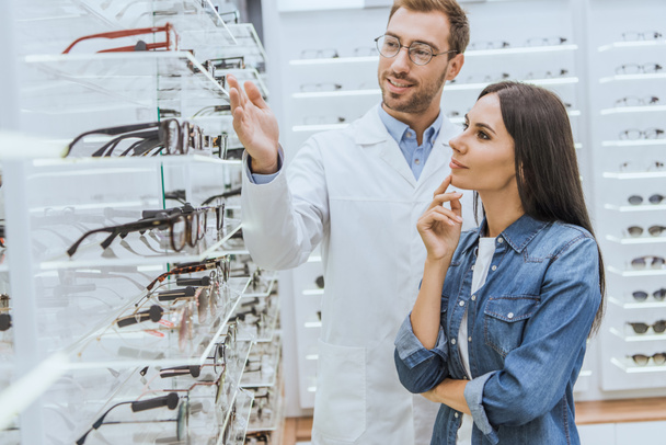 young male optometrist pointing by hand at shelves with eyeglasses to woman in optics  - Photo, Image
