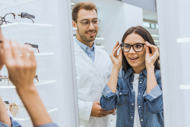 attraktive lächelnde Frau, die eine Brille wählt und in den Spiegel schaut, während der männliche Optiker in der Nähe in der Optik steht  - Foto, Bild