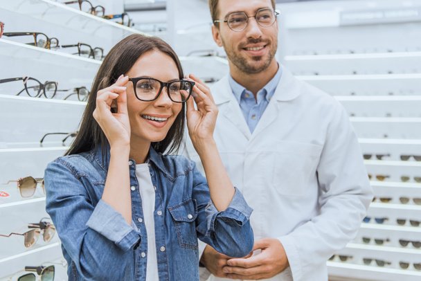 glückliche Frau wählt Brille, während männlicher Augenarzt in der Nähe in Augenarztpraxis steht - Foto, Bild