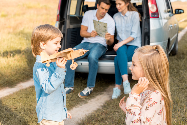 happy little kids playing with toy plane while parents navigating with map and sitting in car trunk in field - Photo, Image