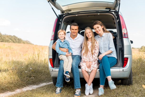happy young family sitting in car trunk and looking at camera in field - Foto, afbeelding
