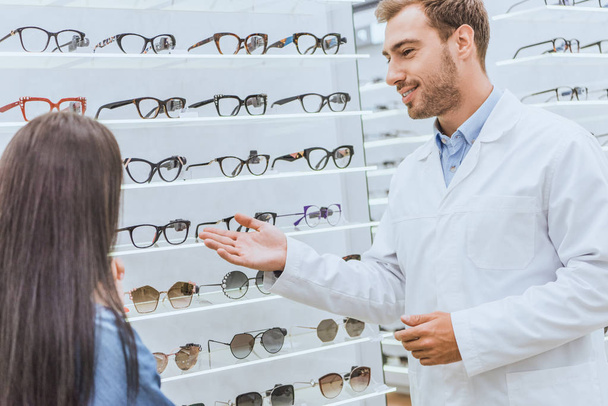 Lächelnder Augenarzt zeigt mit der Hand auf Regale mit Brille auf Frau in Optik - Foto, Bild