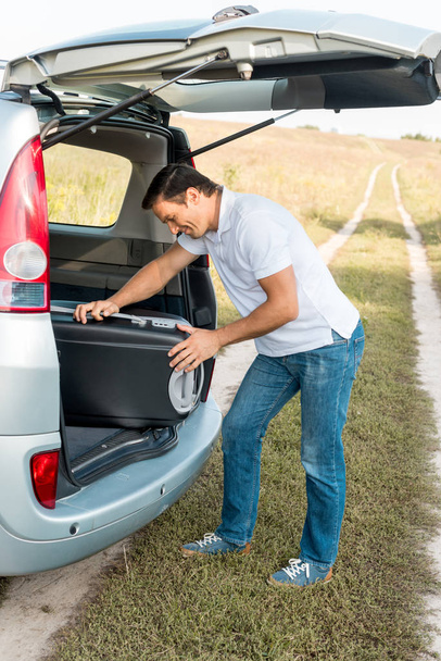 happy man packing suitcase into luggage boot of car in field - Fotó, kép