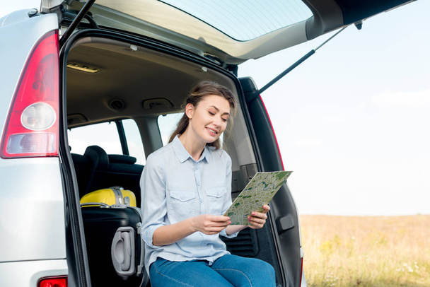 smiling adult woman sitting in car trunk and looking at map in field - Zdjęcie, obraz