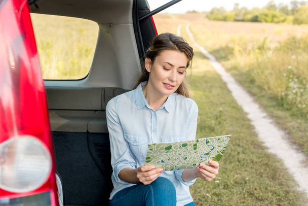 beautiful adult woman sitting in car trunk and looking at map in field - Foto, afbeelding