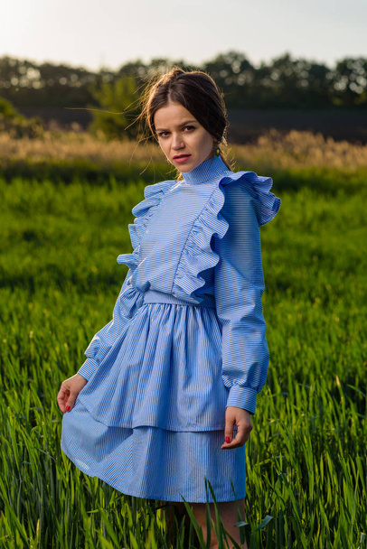 Young woman in blue striped dress is looking towards the camera while standing in the field of green wheat at a spring afternoon during sunny weather. She is holding her dress with one hand. Her hair is fluttering in the wind. - Foto, Imagen