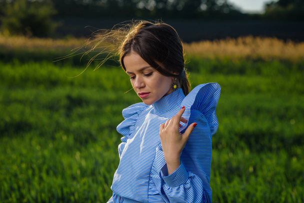 Young woman in blue and white striped dress is looking down while standing in the field of green wheat at a spring afternoon during sunny weather. She is holding one hand at her shoulder. Her hair is fluttering in the wind. - Фото, зображення