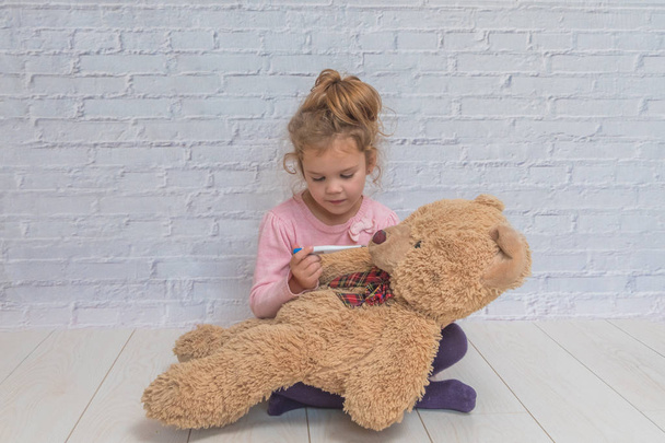 the girl, child measures the temperature of a toy bear, against a white brick wall - Fotoğraf, Görsel