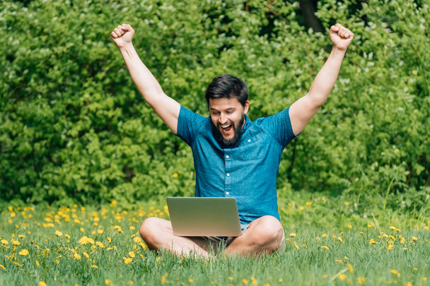 Усміхнений молодий чоловік сидить на зеленій траві з піднятими руками вгору і з ноутбуком на схрещених ногах
 - Фото, зображення