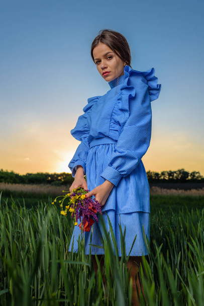 Fiatal nő a kék-fehér csíkos ruha a kezében egy csokor nyári virágok és két kézzel állva a mezőben zöld búza: a tavaszi délután, napsütéses időben, és szeretnének a kamera. A nap beállítást, háta mögött. - Fotó, kép