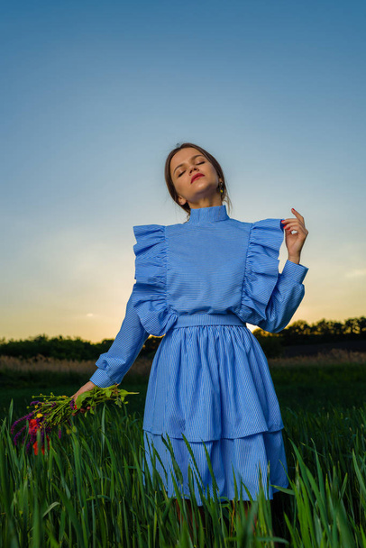 Mujer joven en vestido a rayas azul y blanco está sosteniendo un ramo de flores de verano con una mano mientras está de pie en el campo de trigo verde en una tarde de primavera durante el atardecer. Chica parece que ella está soñando con algo con una de sus manos levantadas
  - Foto, Imagen