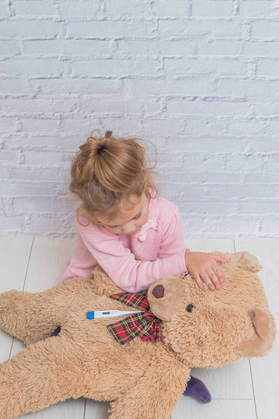 the girl, child measures the temperature of a toy bear, against a white brick wall - Foto, Imagen