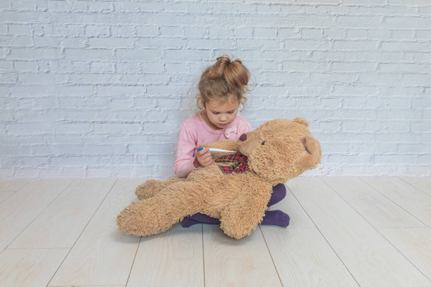 the girl, child measures the temperature of a toy bear, against a white brick wall - Фото, изображение