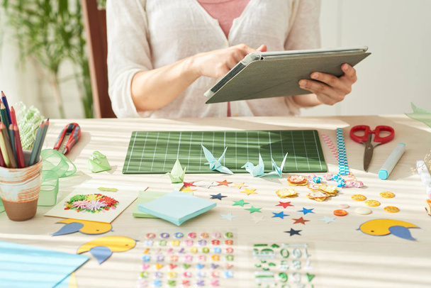 Jonge vrouw zit op houten tafel en met behulp van digitale tablet terwijl het nemen van onderbreking van het maken van origami kranen, decoratieve voorwerpen en instrumenten op de voorgrond - Foto, afbeelding