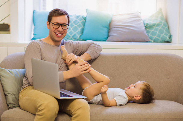 The smiling young father sitting with laptop on a sofa in the room near his cute little son and they playing - Фото, изображение