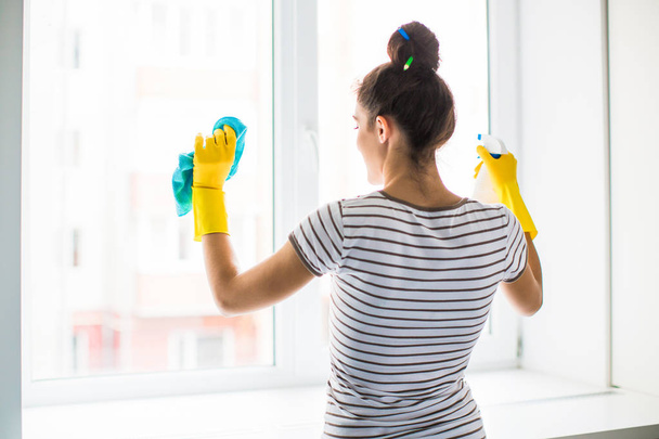 Achteraanzicht van de jonge vrouw die het venster met doek en venster schoner te wassen in de kamer - Foto, afbeelding