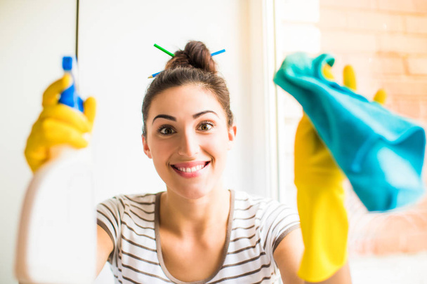 Portrait of the young smiling woman who sitting near the window in the room and holding rag and window cleaner in hands in gloves - Foto, afbeelding