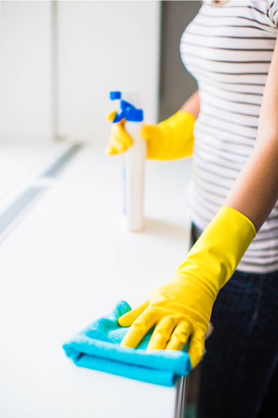 Close-up the young woman in gloves holding rag and window cleaner in hands near the window indoors - Foto, afbeelding