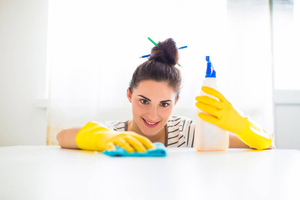 Portrait of the young smiling woman in yellow gloves who holding rag and detergent and washing table in the room - Fotografie, Obrázek