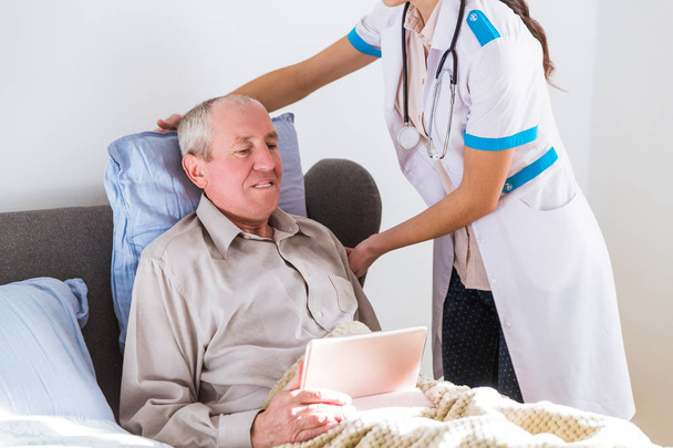 The old, ill, joyful man lying on a bed and holding the tablet in hands and the young doctor standing near him and fixing a pillow to him - Photo, Image