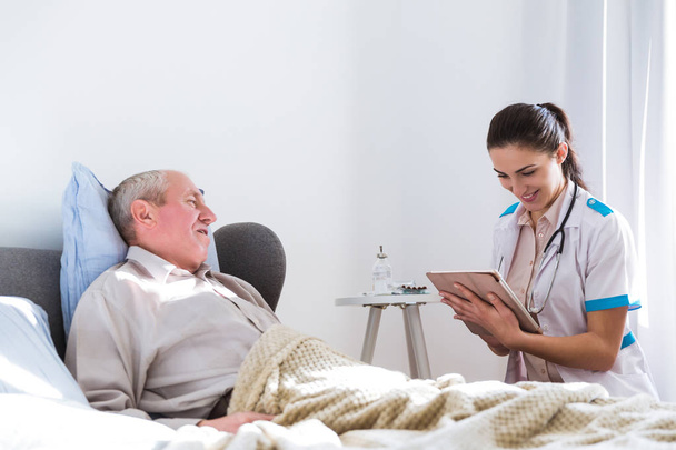 Старий, хворий чоловік лежить на ліжку в кімнаті, дивлячись на молодого лікаря, який сидить поруч з ним і дивиться на планшет
 - Фото, зображення