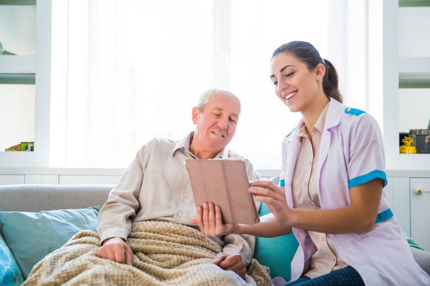 Yaşlı, hasta adam ve onlar evde tablet vasıl seyir yakın oturan genç gülümseyen doktor - Fotoğraf, Görsel